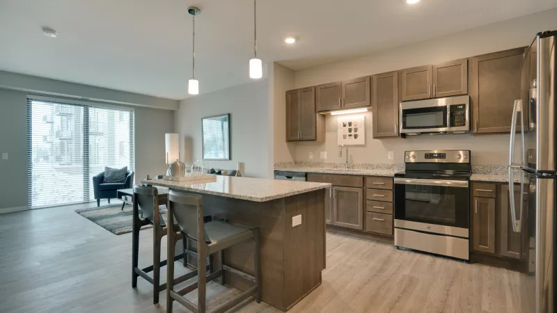 Modern open-plan kitchen with light brown cabinets, stainless steel appliances, an island with bar stools, and an adjacent living room.