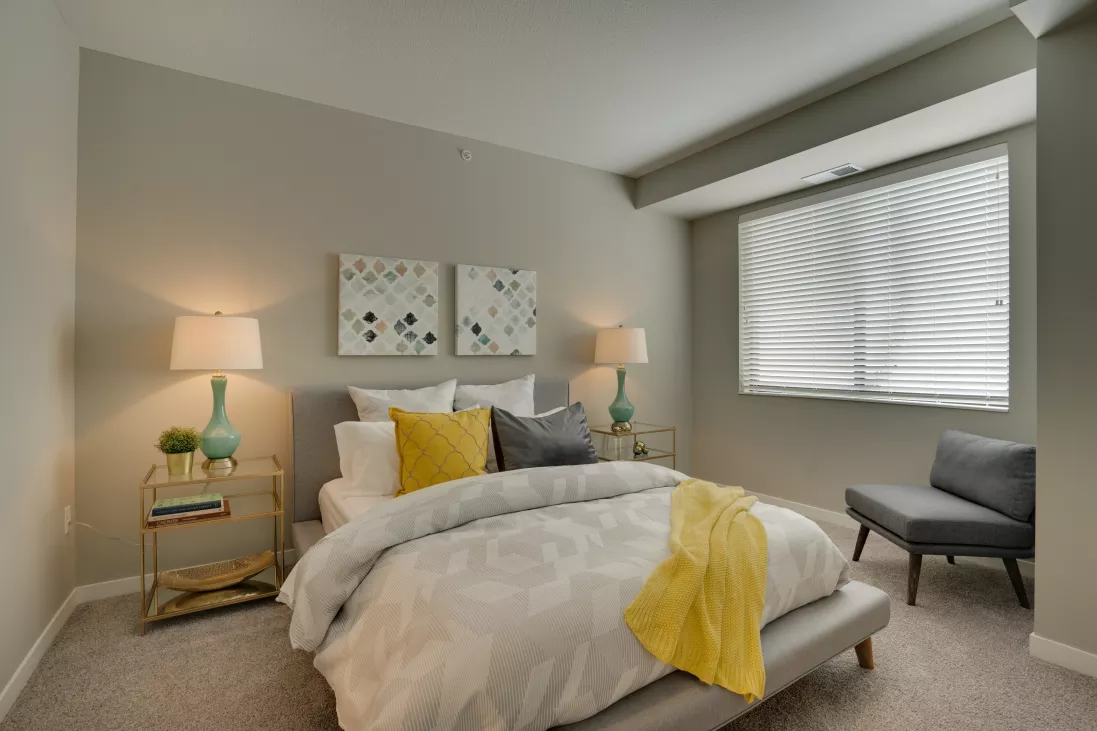 Modern bedroom with gray bed, yellow throw, turquoise lamps, abstract art, and gray armchair by a window.