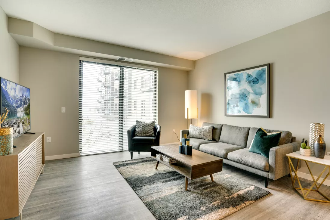 Modern living room with a grey sofa, wooden coffee table, armchair, TV, and abstract art, facing large sliding glass doors.