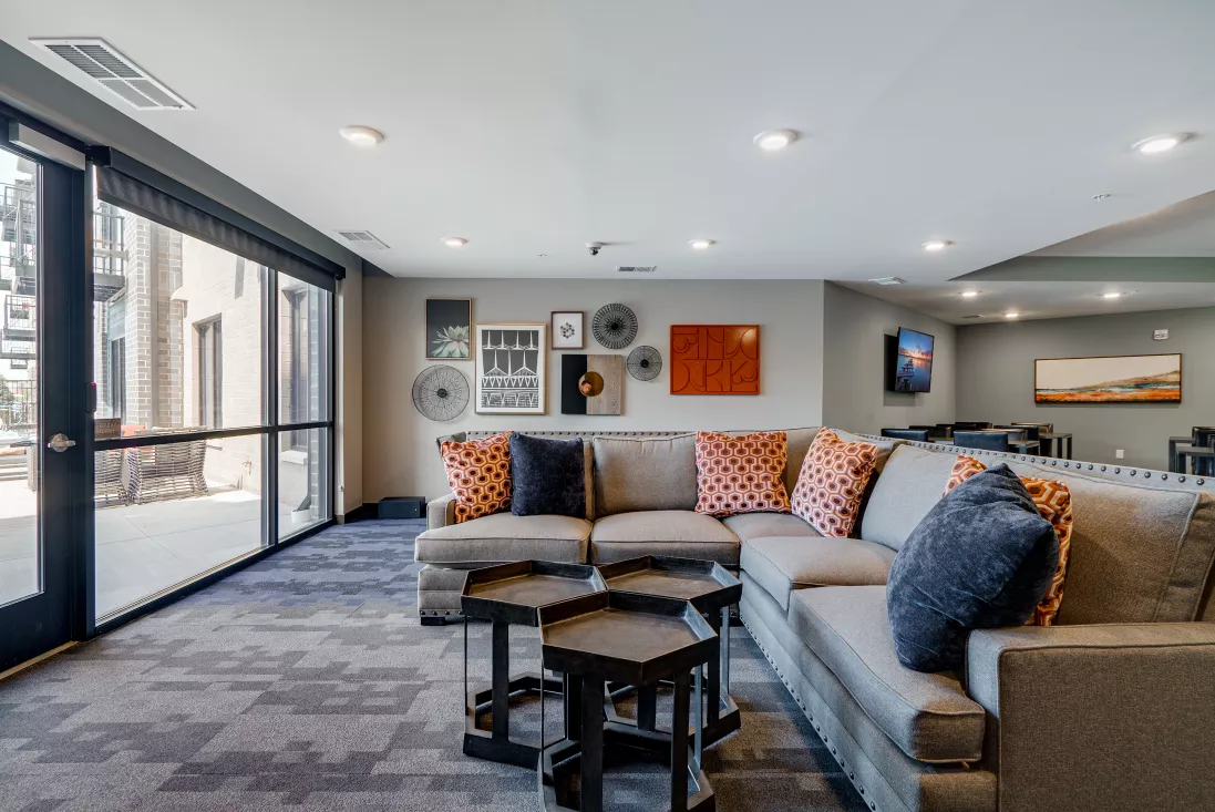Modern lounge with grey sectional sofa, orange and blue pillows, hexagonal tables, wall art, and large windows.
